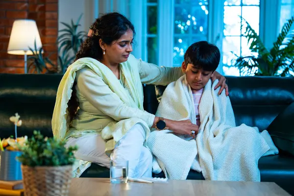 Hintli anne oğlunun ateşini kontrol ederken kanepenin üzerinde oturuyor, sağlığıyla ilgileniyor, sevgisini, sevgisini, sevgisini, aile bağlarını, evde aile yaşam tarzını, günlük rutini besliyor.