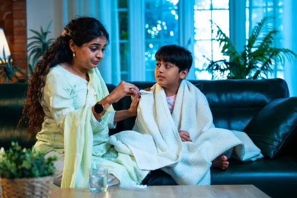 Hintli anne oğlunun ateşini kontrol ederken kanepenin üzerinde oturuyor, sağlığıyla ilgileniyor, sevgisini, sevgisini, sevgisini, aile bağlarını, evde aile yaşam tarzını, günlük rutini besliyor.