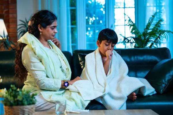 Hintli anne oğlunun ateşini kontrol ederken kanepenin üzerinde oturuyor, sağlığıyla ilgileniyor, sevgisini, sevgisini, sevgisini, aile bağlarını, evde aile yaşam tarzını, günlük rutini besliyor.