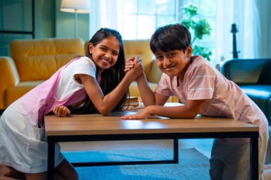 Hintli küçük kardeşler evde bilek güreşi oynuyorlar, Asyalı küçük çocuklar oturma odasında eğlenceli çocukluk aktivitelerinin keyfini çıkarıyorlar, aile bağı, yaşam tarzı, mutluluk.