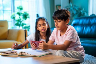 Hintli küçük kardeş evde akıllı telefon ders kitabı ve not defteri kullanarak çalışıyor, Asyalı küçük kardeşler modern oturma odasında birlikte öğreniyor, aile ile eğitim aktivitesi yapıyor.