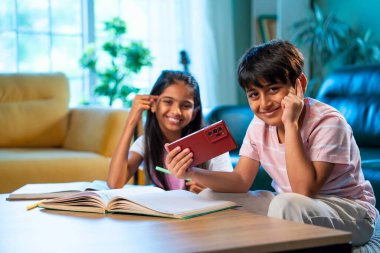 Hintli küçük kardeş evde akıllı telefon ders kitabı ve not defteri kullanarak çalışıyor, Asyalı küçük kardeşler modern oturma odasında birlikte öğreniyor, aile ile eğitim aktivitesi yapıyor.