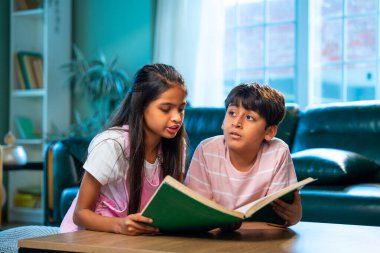 Hintli küçük kardeş evde birlikte kitap okuyor, Asyalı küçük kardeşler öğreniyor, modern oturma odasındaki eğitim aktivitelerine odaklanıyor, aile içinde çocukluk gelişimi.