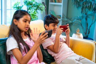 Hintli küçük Asyalı çocuklar evde birlikte mobil oyunlar oynuyorlar, küçük kardeş kardeşler akıllı telefon cihazlarında eğlenceli çocukluk aktivitelerinin tadını çıkarıyorlar, aile içinde kaliteli zaman geçiriyorlar.