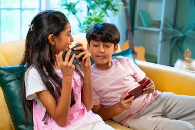 Hintli küçük Asyalı çocuklar evde birlikte mobil oyunlar oynuyorlar, küçük kardeş kardeşler akıllı telefon cihazlarında eğlenceli çocukluk aktivitelerinin tadını çıkarıyorlar, aile içinde kaliteli zaman geçiriyorlar.