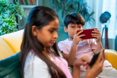 Hintli küçük Asyalı çocuklar evde birlikte mobil oyunlar oynuyorlar, küçük kardeş kardeşler akıllı telefon cihazlarında eğlenceli çocukluk aktivitelerinin tadını çıkarıyorlar, aile içinde kaliteli zaman geçiriyorlar.