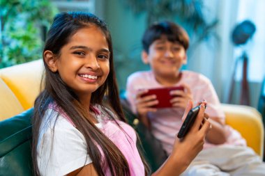 Hintli küçük Asyalı çocuklar evde birlikte mobil oyunlar oynuyorlar, küçük kardeş kardeşler akıllı telefon cihazlarında eğlenceli çocukluk aktivitelerinin tadını çıkarıyorlar, aile içinde kaliteli zaman geçiriyorlar.