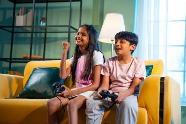 Hintli Asyalı küçük çocuklar evde joystick kontrolörüyle video oyunu oynuyorlar, evde kardeşçe eğlenceli vakit geçiriyorlar, çocukluk eğlenceleri, modern yaşam tarzı dijital eğlence.