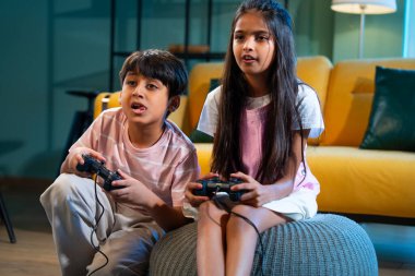 Hintli Asyalı küçük çocuklar evde joystick kontrolörüyle video oyunu oynuyorlar, evde kardeşçe eğlenceli vakit geçiriyorlar, çocukluk eğlenceleri, modern yaşam tarzı dijital eğlence.