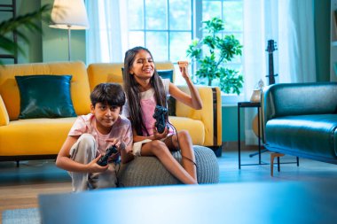 Hintli Asyalı küçük çocuklar evde joystick kontrolörüyle video oyunu oynuyorlar, evde kardeşçe eğlenceli vakit geçiriyorlar, çocukluk eğlenceleri, modern yaşam tarzı dijital eğlence.