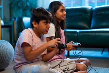 Hintli Asyalı küçük çocuklar evde joystick kontrolörüyle video oyunu oynuyorlar, evde kardeşçe eğlenceli vakit geçiriyorlar, çocukluk eğlenceleri, modern yaşam tarzı dijital eğlence.