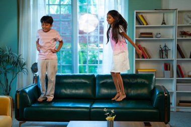 Hintli Asyalı küçük çocuklar mutlu bir şekilde modern ev oturma odasında kanepede zıplayıp, kardeş olarak kaliteli vakit geçirmenin, neşeli çocukluk aktivitelerinin, neşeli aile bağlarının, kapalı alanda eğlencenin tadını çıkarıyorlar.