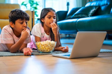 Hintli Asyalı küçük kardeş diziler izlerken dizüstü bilgisayar başında patlamış mısır yiyor, modern oturma odasında halının üzerinde oturuyor, eğlence, eğlence aktivitesi, bağ kurma, mutlu çocukluk.