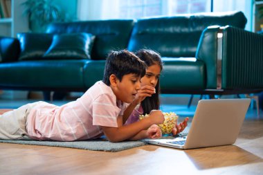 Hintli Asyalı küçük kardeş diziler izlerken dizüstü bilgisayar başında patlamış mısır yiyor, modern oturma odasında halının üzerinde oturuyor, eğlence, eğlence aktivitesi, bağ kurma, mutlu çocukluk.