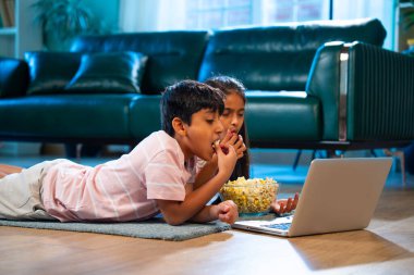 Hintli Asyalı küçük kardeş diziler izlerken dizüstü bilgisayar başında patlamış mısır yiyor, modern oturma odasında halının üzerinde oturuyor, eğlence, eğlence aktivitesi, bağ kurma, mutlu çocukluk.