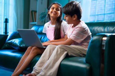 Hintli küçük kardeş dizüstü bilgisayar ve akıllı telefon kullanırken modern ev içlerindeki kanepede oturuyor, Asyalı kardeş kardeş çocuklar dijital yaşam tarzının, bağlanmanın, kapalı alandaki çocukluk eğlencesinin tadını çıkarıyorlar.