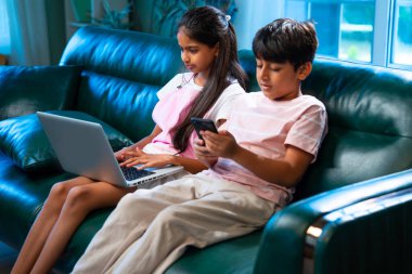 Hintli küçük kardeş dizüstü bilgisayar ve akıllı telefon kullanırken modern ev içlerindeki kanepede oturuyor, Asyalı kardeş kardeş çocuklar dijital yaşam tarzının, bağlanmanın, kapalı alandaki çocukluk eğlencesinin tadını çıkarıyorlar.