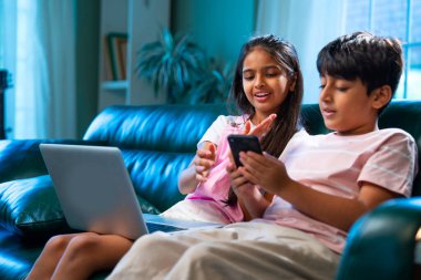 Hintli küçük kardeş dizüstü bilgisayar ve akıllı telefon kullanırken modern ev içlerindeki kanepede oturuyor, Asyalı kardeş kardeş çocuklar dijital yaşam tarzının, bağlanmanın, kapalı alandaki çocukluk eğlencesinin tadını çıkarıyorlar.