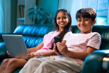 Hintli küçük kardeş dizüstü bilgisayar ve akıllı telefon kullanırken modern ev içlerindeki kanepede oturuyor, Asyalı kardeş kardeş çocuklar dijital yaşam tarzının, bağlanmanın, kapalı alandaki çocukluk eğlencesinin tadını çıkarıyorlar.