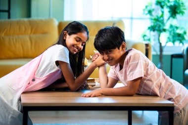 Hintli küçük kardeşler evde bilek güreşi oynuyorlar, Asyalı küçük çocuklar oturma odasında eğlenceli çocukluk aktivitelerinin keyfini çıkarıyorlar, aile bağı, yaşam tarzı, mutluluk.