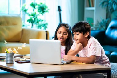 Hintli küçük kardeş evde dizüstü bilgisayarla ders çalışıyor, evde not defteri var, Asyalı küçük çocuklar birlikte öğreniyorlar modern oturma odasında bilgisayar teknolojisi, eğitim ve ev ödevi kullanıyorlar.