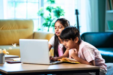 Hintli küçük kardeş evde dizüstü bilgisayarla ders çalışıyor, evde not defteri var, Asyalı küçük çocuklar birlikte öğreniyorlar modern oturma odasında bilgisayar teknolojisi, eğitim ve ev ödevi kullanıyorlar.