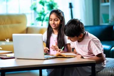 Hintli küçük kardeş evde dizüstü bilgisayarla ders çalışıyor, evde not defteri var, Asyalı küçük çocuklar birlikte öğreniyorlar modern oturma odasında bilgisayar teknolojisi, eğitim ve ev ödevi kullanıyorlar.