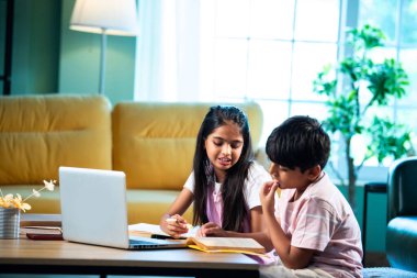 Hintli küçük kardeş evde dizüstü bilgisayarla ders çalışıyor, evde not defteri var, Asyalı küçük çocuklar birlikte öğreniyorlar modern oturma odasında bilgisayar teknolojisi, eğitim ve ev ödevi kullanıyorlar.