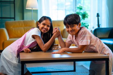 Hintli küçük kardeşler evde bilek güreşi oynuyorlar, Asyalı küçük çocuklar oturma odasında eğlenceli çocukluk aktivitelerinin keyfini çıkarıyorlar, aile bağı, yaşam tarzı, mutluluk.