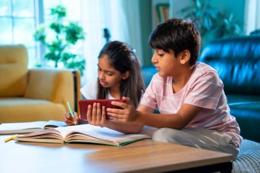 Hintli küçük kardeş evde akıllı telefon ders kitabı ve not defteri kullanarak çalışıyor, Asyalı küçük kardeşler modern oturma odasında birlikte öğreniyor, aile ile eğitim aktivitesi yapıyor.