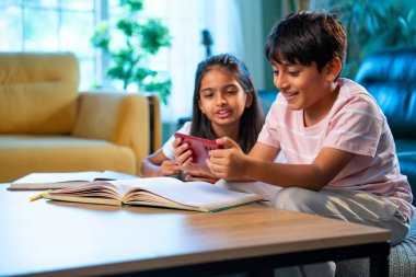 Hintli küçük kardeş evde akıllı telefon ders kitabı ve not defteri kullanarak çalışıyor, Asyalı küçük kardeşler modern oturma odasında birlikte öğreniyor, aile ile eğitim aktivitesi yapıyor.