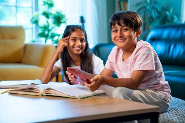 Hintli küçük kardeş evde akıllı telefon ders kitabı ve not defteri kullanarak çalışıyor, Asyalı küçük kardeşler modern oturma odasında birlikte öğreniyor, aile ile eğitim aktivitesi yapıyor.