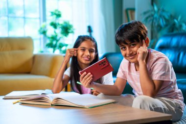Hintli küçük kardeş evde akıllı telefon ders kitabı ve not defteri kullanarak çalışıyor, Asyalı küçük kardeşler modern oturma odasında birlikte öğreniyor, aile ile eğitim aktivitesi yapıyor.
