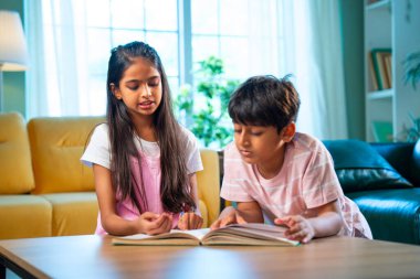 Hintli küçük kardeş evde birlikte kitap okuyor, Asyalı küçük kardeşler öğreniyor, modern oturma odasındaki eğitim aktivitelerine odaklanıyor, aile içinde çocukluk gelişimi.