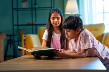 Hintli küçük kardeş evde birlikte kitap okuyor, Asyalı küçük kardeşler öğreniyor, modern oturma odasındaki eğitim aktivitelerine odaklanıyor, aile içinde çocukluk gelişimi.