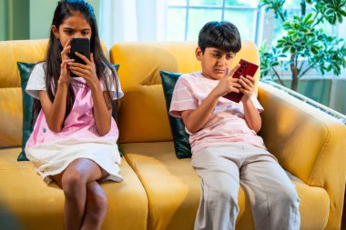 Hintli küçük Asyalı çocuklar evde birlikte mobil oyunlar oynuyorlar, küçük kardeş kardeşler akıllı telefon cihazlarında eğlenceli çocukluk aktivitelerinin tadını çıkarıyorlar, aile içinde kaliteli zaman geçiriyorlar.