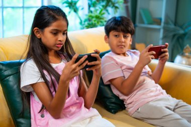 Hintli küçük Asyalı çocuklar evde birlikte mobil oyunlar oynuyorlar, küçük kardeş kardeşler akıllı telefon cihazlarında eğlenceli çocukluk aktivitelerinin tadını çıkarıyorlar, aile içinde kaliteli zaman geçiriyorlar.