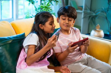 Hintli küçük Asyalı çocuklar evde birlikte mobil oyunlar oynuyorlar, küçük kardeş kardeşler akıllı telefon cihazlarında eğlenceli çocukluk aktivitelerinin tadını çıkarıyorlar, aile içinde kaliteli zaman geçiriyorlar.