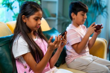 Hintli küçük Asyalı çocuklar evde birlikte mobil oyunlar oynuyorlar, küçük kardeş kardeşler akıllı telefon cihazlarında eğlenceli çocukluk aktivitelerinin tadını çıkarıyorlar, aile içinde kaliteli zaman geçiriyorlar.