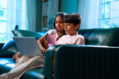 Hintli küçük kardeş dizüstü bilgisayar ve akıllı telefon kullanırken modern ev içlerindeki kanepede oturuyor, Asyalı kardeş kardeş çocuklar dijital yaşam tarzının, bağlanmanın, kapalı alandaki çocukluk eğlencesinin tadını çıkarıyorlar.