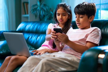 Hintli küçük kardeş dizüstü bilgisayar ve akıllı telefon kullanırken modern ev içlerindeki kanepede oturuyor, Asyalı kardeş kardeş çocuklar dijital yaşam tarzının, bağlanmanın, kapalı alandaki çocukluk eğlencesinin tadını çıkarıyorlar.