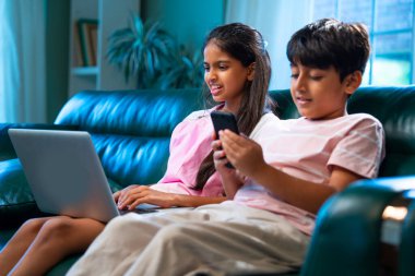 Hintli küçük kardeş dizüstü bilgisayar ve akıllı telefon kullanırken modern ev içlerindeki kanepede oturuyor, Asyalı kardeş kardeş çocuklar dijital yaşam tarzının, bağlanmanın, kapalı alandaki çocukluk eğlencesinin tadını çıkarıyorlar.