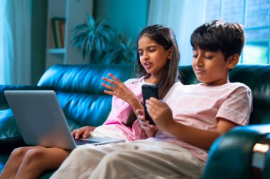 Hintli küçük kardeş dizüstü bilgisayar ve akıllı telefon kullanırken modern ev içlerindeki kanepede oturuyor, Asyalı kardeş kardeş çocuklar dijital yaşam tarzının, bağlanmanın, kapalı alandaki çocukluk eğlencesinin tadını çıkarıyorlar.