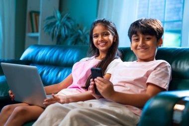 Hintli küçük kardeş dizüstü bilgisayar ve akıllı telefon kullanırken modern ev içlerindeki kanepede oturuyor, Asyalı kardeş kardeş çocuklar dijital yaşam tarzının, bağlanmanın, kapalı alandaki çocukluk eğlencesinin tadını çıkarıyorlar.