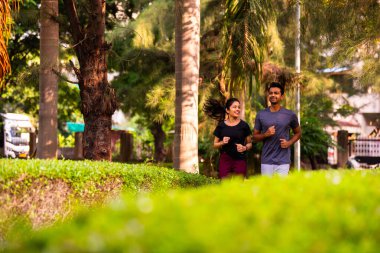 Hintli genç bir çift parkta birlikte koşuyor sağlıklı sabah rutinlerinin ve spor odaklı günün başlangıcında ağaçlarla çevrili temiz havayı soluyarak kaliteli zaman geçiriyorlar.