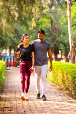 Hintli güzel genç çift sabah vakti parkta yürüyorlar birlikte huzurlu vakit geçiriyorlar, gülüyorlar, konuşuyorlar, bağ kuruyorlar, doğayı deneyimliyorlar, sağlıklı ve aktif bir yaşam sürüyorlar.