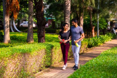 Hintli güzel genç çift sabah vakti parkta yürüyorlar birlikte huzurlu vakit geçiriyorlar, gülüyorlar, konuşuyorlar, bağ kuruyorlar, doğayı deneyimliyorlar, sağlıklı ve aktif bir yaşam sürüyorlar.
