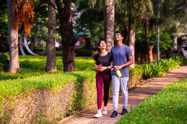 Hintli güzel genç çift sabah vakti parkta yürüyorlar birlikte huzurlu vakit geçiriyorlar, gülüyorlar, konuşuyorlar, bağ kuruyorlar, doğayı deneyimliyorlar, sağlıklı ve aktif bir yaşam sürüyorlar.