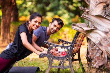 Hintli güzel genç çift sabah spor salonunda parkta şınav çekiyorlar. Dayanıklılıklarını, takım çalışmalarını ve sağlıklı yaşam tarzlarını gösteriyorlar. Güneş ışığının, temiz havanın ve yeşilliğin tadını çıkarıyorlar.