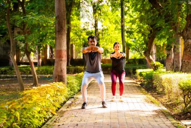 Hintli güzel genç çift güneşli bir günde parkın dışında birlikte çömeliyorlar. Günlük fitness rutinlerinin bir parçası olarak, güç, sağlık, motivasyon ve aktif sağlıklı yaşam tarzı hedeflerine odaklanıyorlar.